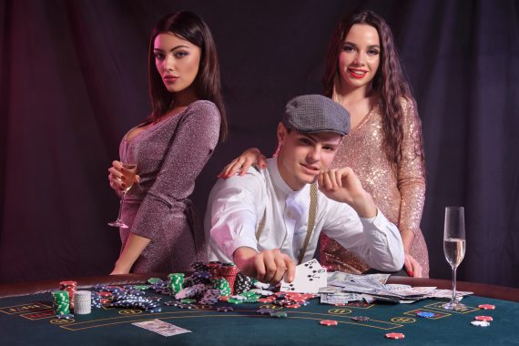 Strategie bonus poker online - Man playing poker at casino sitting at table with stacks of chips, money, cards. Celebrating win with two women. Black background. Close up.